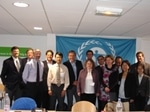 A group of people in business attire stands together, smiling for a photo in a conference room. A blue flag with a white emblem hangs on the wall behind them.