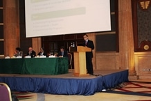 A person stands at a podium giving a presentation in a large room, while five people sit at a table on stage. A projection screen displays information behind them.
