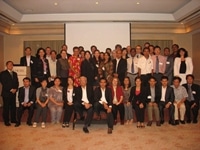 A large group of people in business attire pose together for a group photo in a conference room, with a projector screen in the background.
