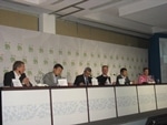 Six people sit at a long conference table on a stage, each with nameplates and microphones, appearing to participate in a panel discussion. The background displays a patterned banner.