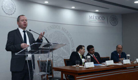 A man speaks at a podium while four men sit at a table in a conference room with a Mexico government emblem on the wall.