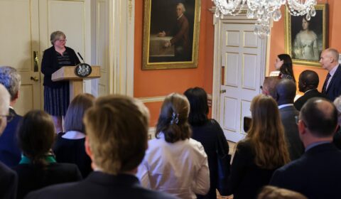 A woman speaks at a podium to a standing audience in an ornate room with chandeliers and paintings on the walls.