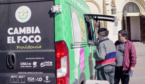 Two people stand next to a green van with the text Cambia el foco Providencia on it, parked near a building entrance. One person talks to someone inside the van through a window.