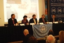 Five people sit at a panel table with microphones, facing an audience. A blue flag with a globe emblem hangs in front. A projection screen and backdrop with logos are visible behind them.