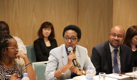 A woman in a striped blazer speaks into a microphone at a conference table, surrounded by colleagues who listen attentively. Bottled water, notebooks, and cups are on the table.