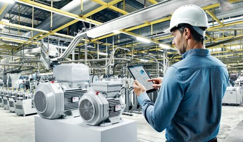 A man in a hard hat uses a tablet to inspect large industrial machinery inside a modern, brightly lit factory filled with equipment and overhead piping.
