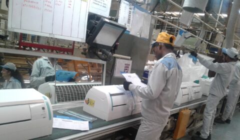 Workers in uniforms operate machinery and process documents on an assembly line in a factory setting. One worker examines papers, while another handles a large plastic bag nearby. Monitors and charts are visible above the line.