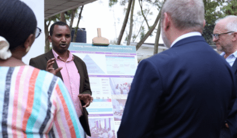 A man presents information on a poster to three people outdoors. The group listens attentively, and trees are visible in the background.