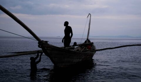 Silhouettes of people on and near a wooden boat in calm water during dusk, with a cloudy sky and distant land visible in the background.