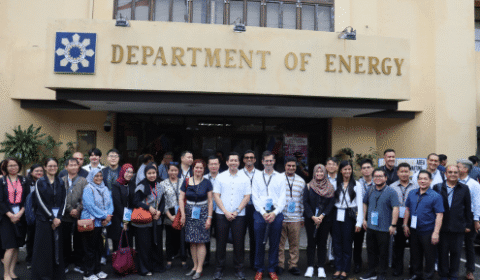 A large group of people stands smiling in front of a building with a Department of Energy sign above the entrance. Some people wear name lanyards and business attire.