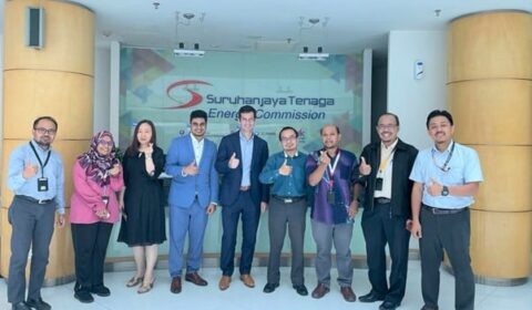 Ten people stand in a row, smiling and posing with thumbs up, in front of a sign that reads Suruhanjaya Tenaga Energy Commission..