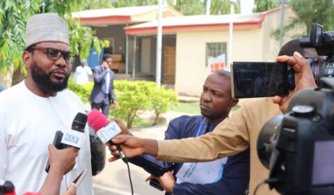 A man in glasses and traditional attire speaks to reporters holding microphones and recording devices outside a building, with trees and another person visible in the background.