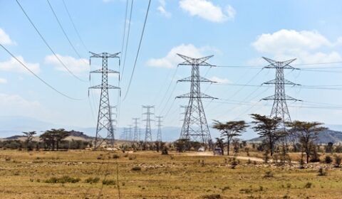 Several tall power transmission towers with electrical wires run across a dry, grassy landscape with scattered trees under a partly cloudy sky.