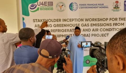 A man speaks to a group of journalists and cameras at a workshop about reducing greenhouse gas emissions, standing in front of a banner with project and sponsor logos.
