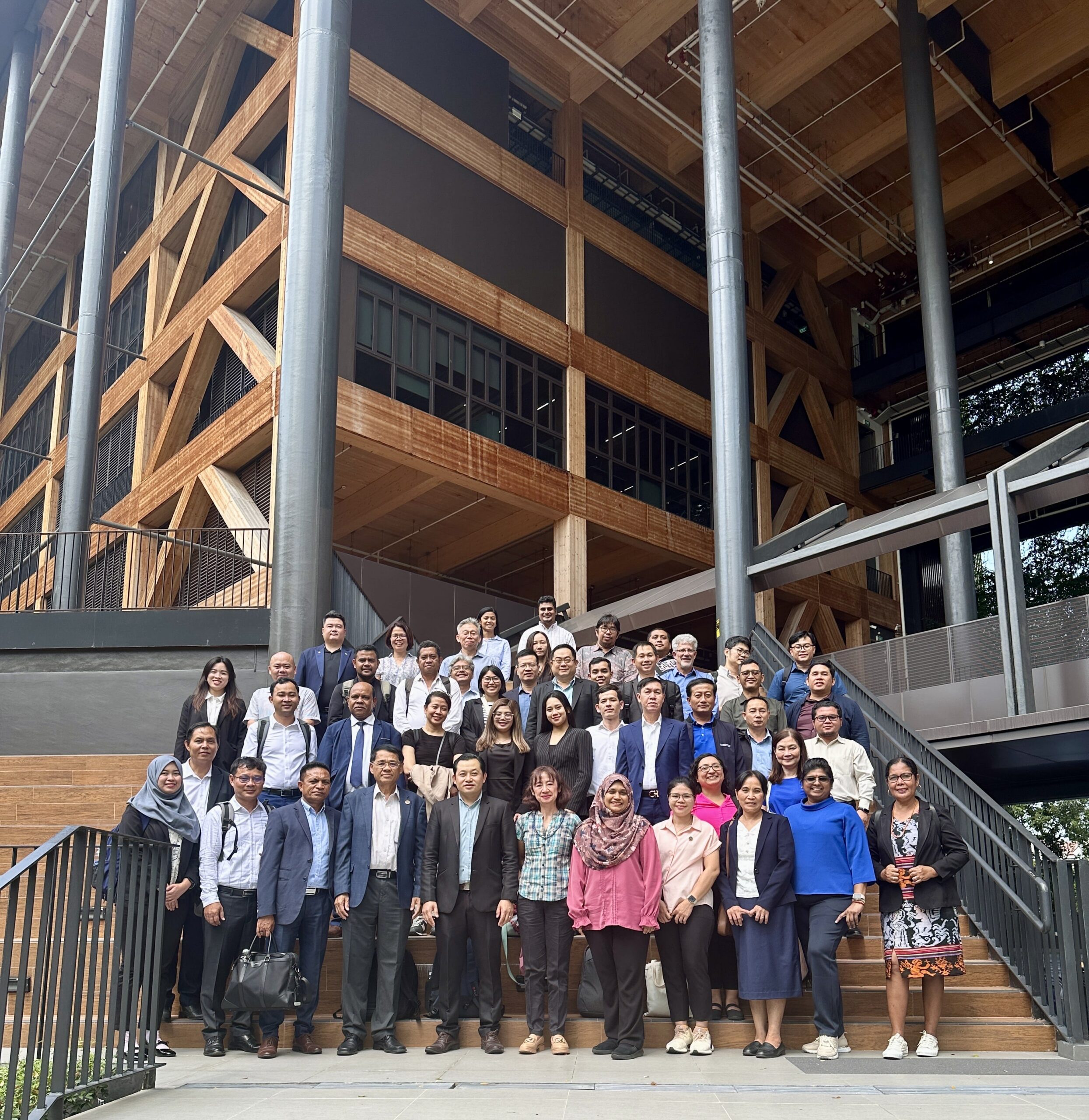 A large group of people pose together on outdoor stairs in front of a modern wooden building, smiling and dressed in business or smart casual attire.