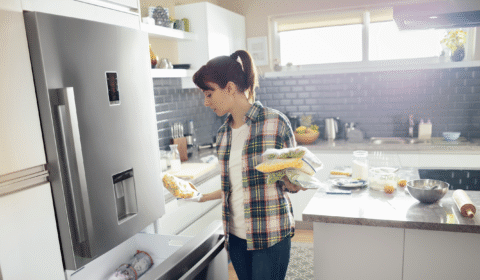 Woman wearing a plaid shirt places frozen food into the freezer drawer of a stainless steel refrigerator in a modern kitchen with gray cabinets and a large window.