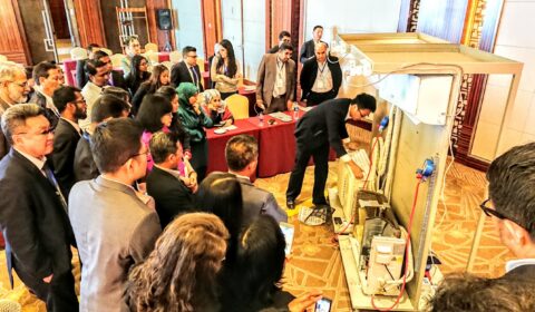 A group of people in formal attire watch a man demonstrating equipment in a conference room. Attendees are gathered closely, some seated, most standing, observing the demonstration with interest.