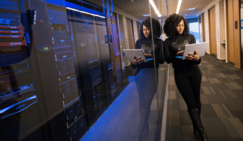 A woman stands in a server room, holding and working on a laptop. She is reflected in the glass wall beside her, surrounded by server racks with blue lighting.