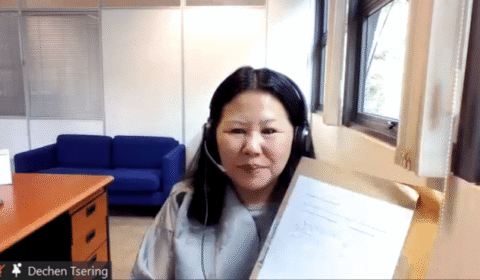 A woman with headphones sits at a desk, holding up a document to the camera during a video call. Her name, Dechen Tsering, appears in the corner of the screen.