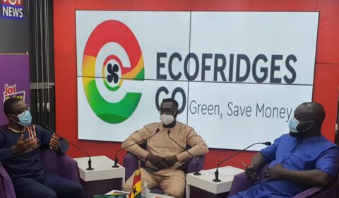Three men in masks sit in a TV studio discussing ECOFRIDGES GO, with the slogan Green, Save Money on a screen behind them. Newspapers and a small Ghana flag are on the table.