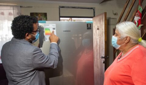 A man in a suit shows an energy efficiency label on a refrigerator to a woman in a pink shirt; both are wearing masks and standing indoors.