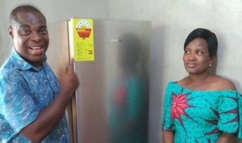 A smiling man points at a refrigerator with a sticker while a woman in a green patterned dress stands beside him, looking on calmly.
