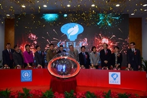 A group of people in formal attire stand behind a red table with a large transparent globe, as confetti falls and fireworks display on a screen behind them. The event appears to be a celebration or ceremony.