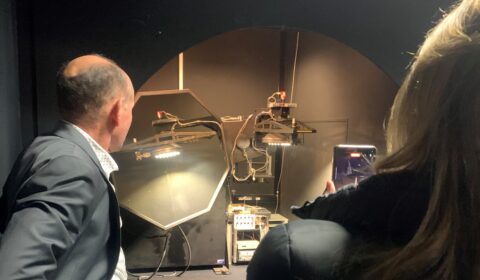 Two people observe a complex machine with lights and wires inside a dark room; one person is taking a photo with a phone.