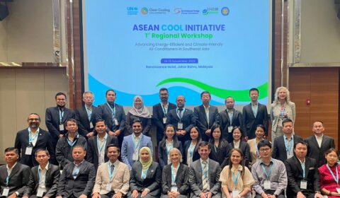 A large group of men and women pose for a photo in business attire at the ASEAN Cool Initiative Regional Workshop, with a conference presentation screen behind them.