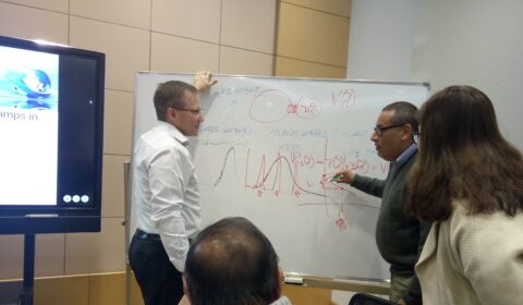 Three people stand at a whiteboard covered in mathematical equations and diagrams, discussing and pointing at the content. A seated person listens nearby, with a large screen visible in the background.