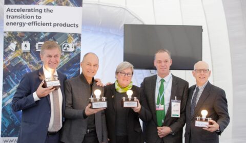Five people in business attire hold trophies with light bulbs. A banner behind them reads Accelerating the transition to energy-efficient products. They are smiling and standing together at an event.