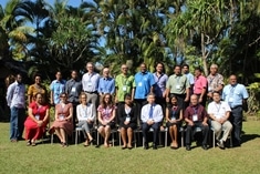 A group of people pose outdoors for a photo, with some seated in front and others standing behind. They are dressed in business casual attire, and tropical trees and plants fill the background.