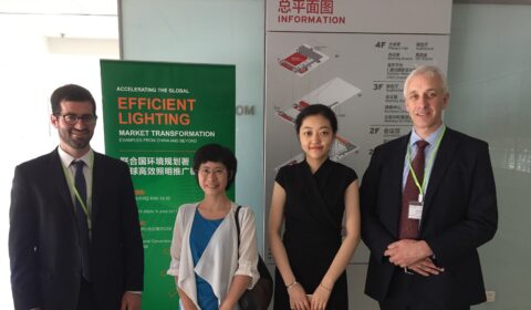 Four people in business attire stand indoors in front of a green Efficient Lighting poster and a wall directory with maps. They are smiling and wearing conference badges or lanyards.