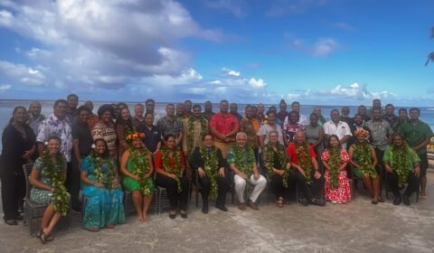 A large group of people pose for a photo outdoors by the ocean, many wearing leis and tropical attire, with blue sky and clouds in the background.