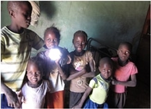 Six children stand together indoors, smiling at the camera. One child in the middle holds a bright, glowing light source, illuminating their faces against a dim, greenish wall background.