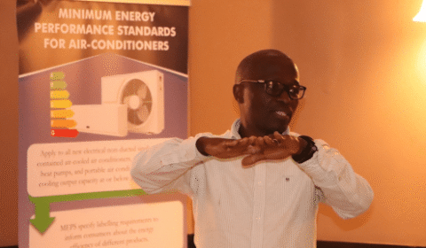 A man in glasses gestures with his hands while standing in front of a sign about minimum energy performance standards for air conditioners.
