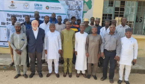 A group of men pose for a photo outside a building, in front of a banner that reads Training Workshop on End-of-life Air Conditioning. Some are in suits, others in traditional attire.