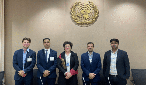Five people in business attire stand in a row, posing for a photo under a large United Nations emblem on a beige wall.