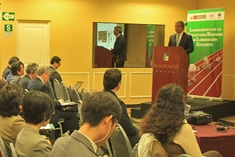 A man speaks at a podium during a conference, with attendees seated and listening. A presentation is projected on the wall behind him.