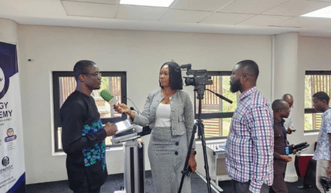 A man stands at a podium speaking, while a woman interviews him with a microphone and another man records with a video camera. Several people are in the background in a bright, modern room.
