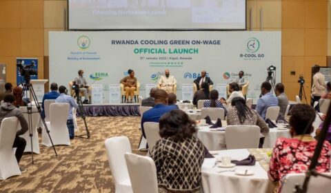 People attend the official launch event for the Rwanda Cooling Green On-Wage initiative in Kigali, with a panel on stage, audience members seated, and cameras recording. Banners and branding are visible in the background.