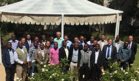 A group of about 25 people stands together outdoors in front of a white canopy, with green bushes and white flowers in the foreground, posing for a group photo in sunny weather.