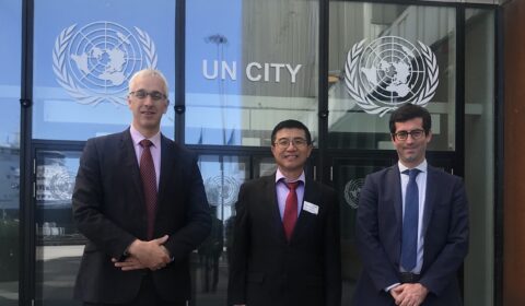 Three men in suits stand outside a building with UN CITY and United Nations logos on glass doors, posing for a photo on a sunny day.
