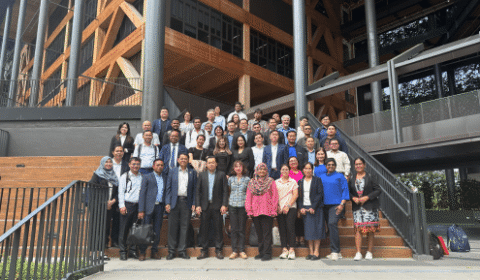 A large group of people pose together on stairs outside a modern building with wood and glass construction, smiling for a group photo.