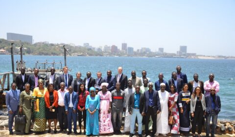 A large group of people, dressed in business and traditional attire, pose for a photo outdoors by the water, with a city skyline visible in the background.