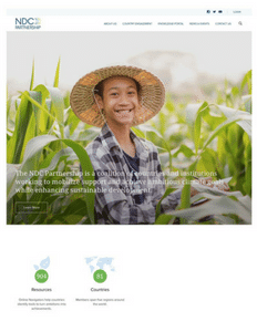 A smiling person wearing a straw hat stands in a green cornfield. The website header and navigation menu are visible above a text overlay about the NDC Partnerships mission. Resources and countries served are shown below.