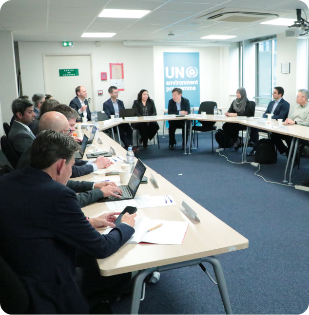 People sit around a U-shaped conference table with laptops and papers. A UN Environment Programme sign is displayed in the background. The meeting appears formal and collaborative.