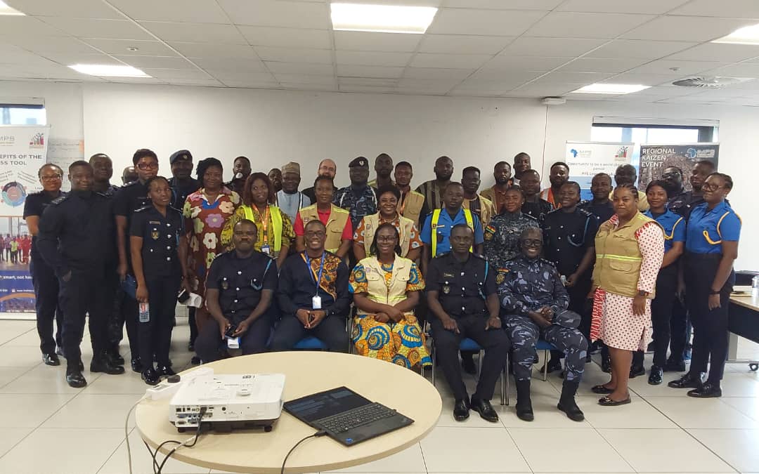 A group of men and women, some in police or security uniforms, pose together in a bright room with banners, a projector, and a laptop in the foreground.