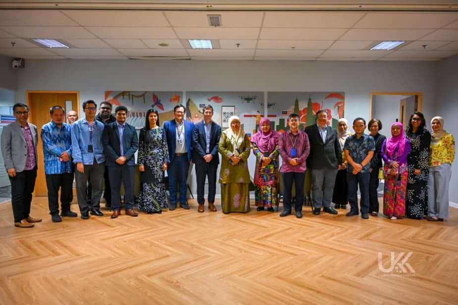 A group of fifteen people, men and women, stand in a row indoors, dressed in business and traditional attire, posing for a group photo in a brightly lit room with artwork on the wall behind them.
