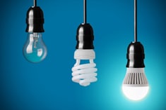 Three hanging light bulbs: an incandescent bulb, a compact fluorescent bulb (CFL), and a glowing LED bulb, against a blue background.